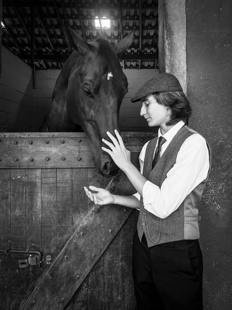 Jovem de colete e boina acariciando um cavalo na baia