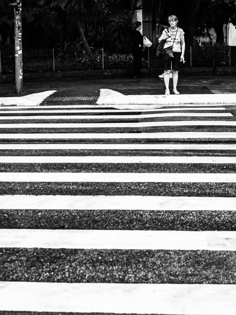 Faixa de pedestres em primeiro plano e uma pessoa esperando para atravessar